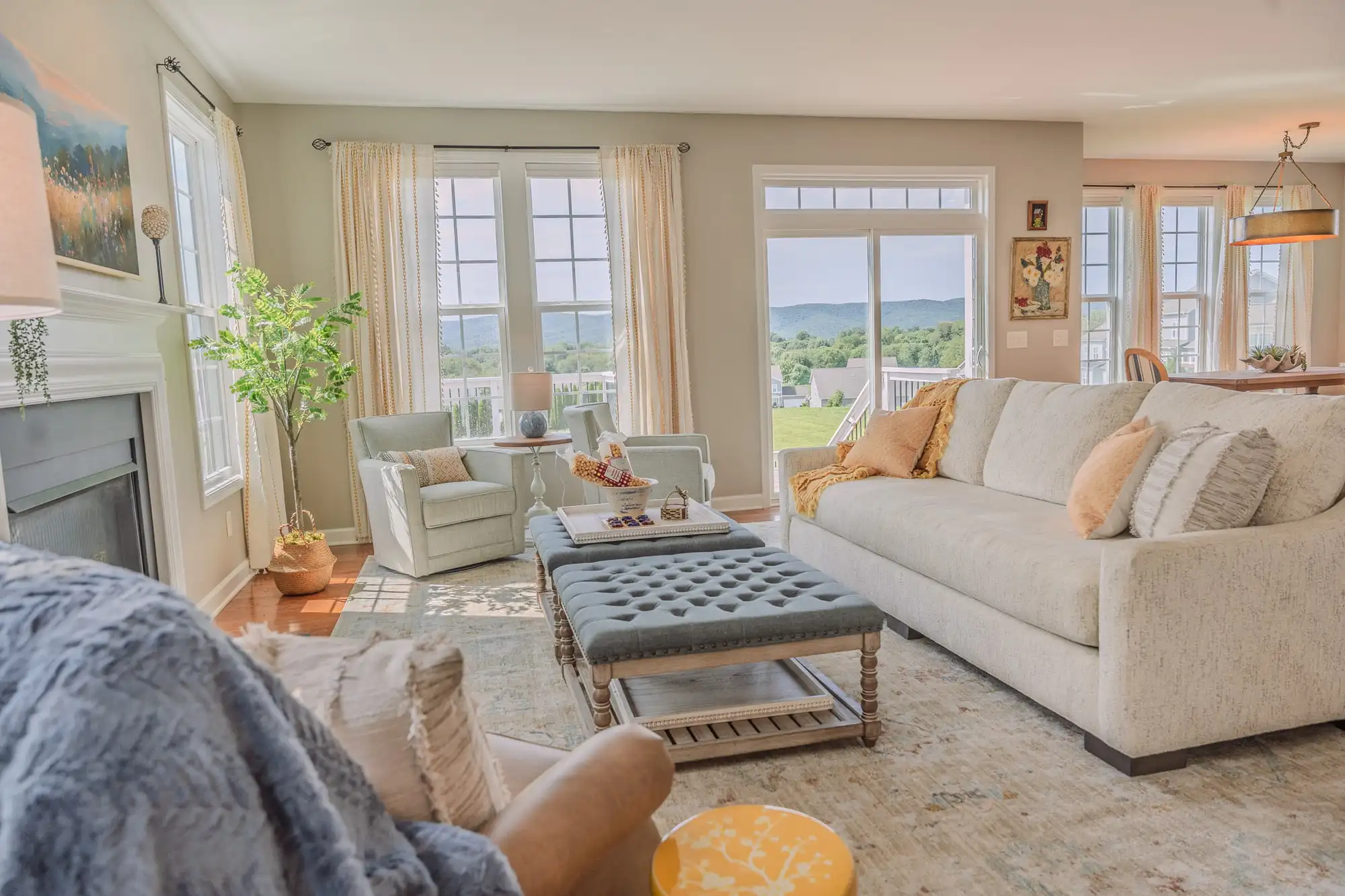 Living room with a pale sofa, blue tufted ottomans, yellow cushions, and windows overlooking green hills