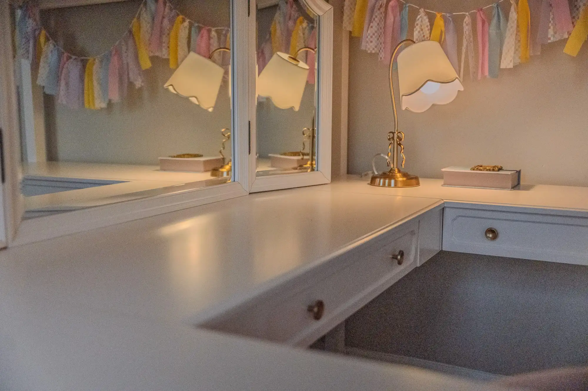 Desk beneath a loft bed with two mirrors, a pastel garland, and a scalloped lamp