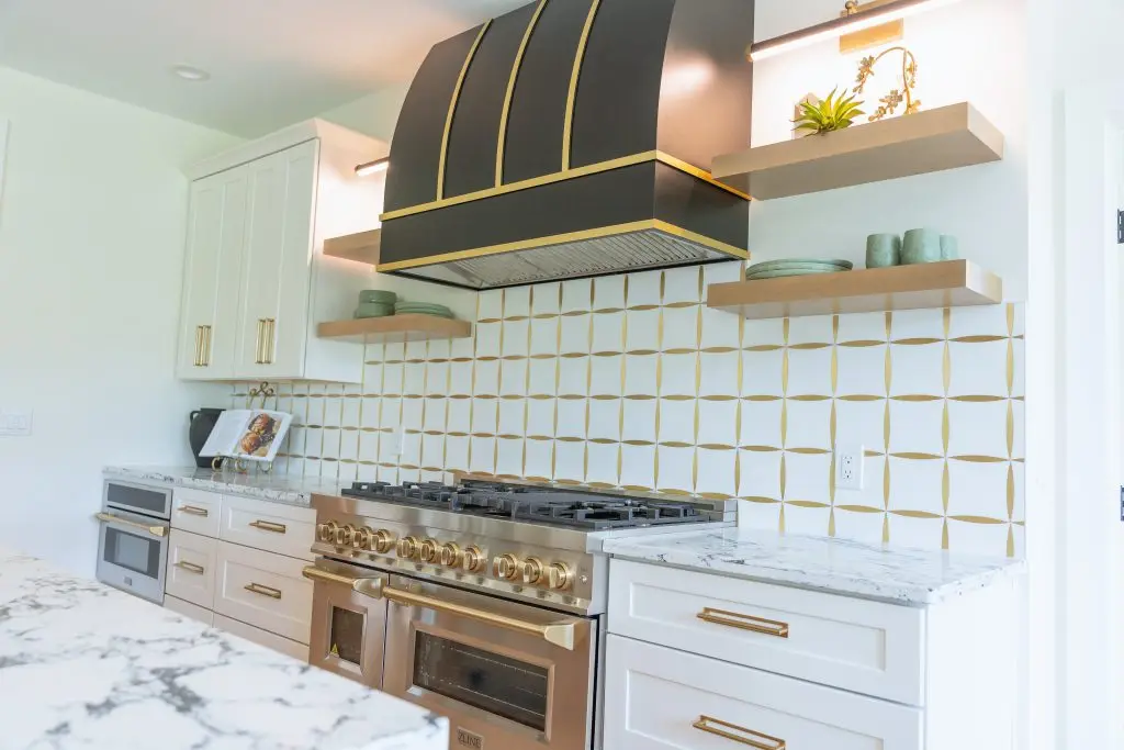 Range beneath a dark hood with brass trim, patterned tile, and open wood shelves