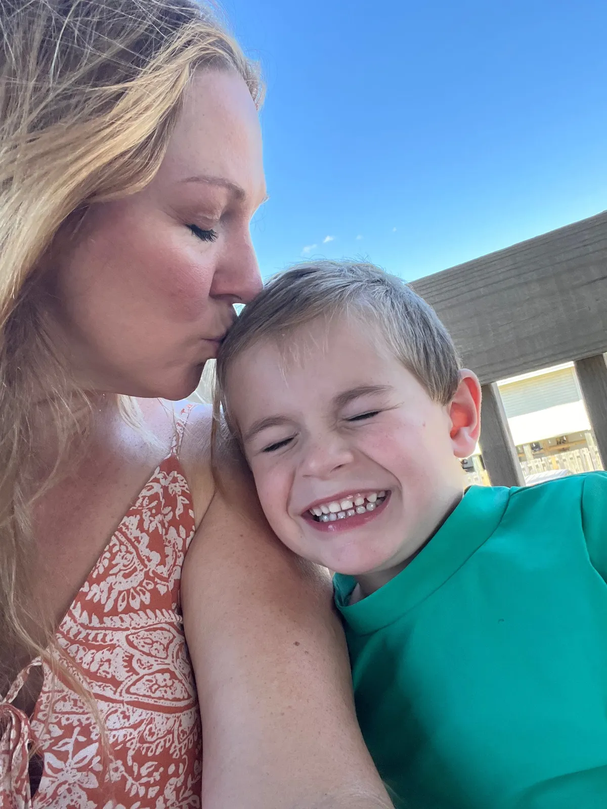 Woman kissing a smiling child on the cheek outdoors under a clear blue sky, sunlight on their faces.