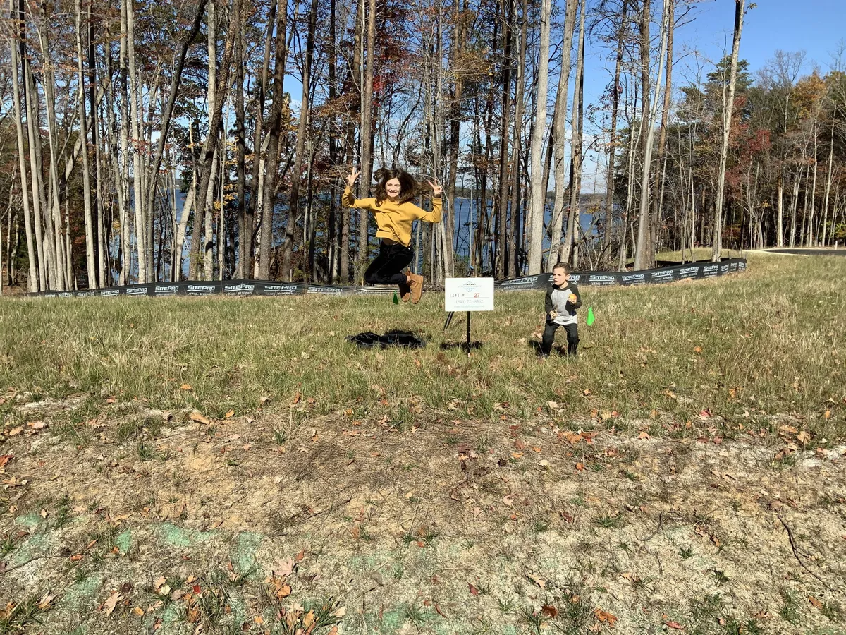 Girl in a mustard sweater jumps with peace signs in a grassy park, autumn trees and a lake in the background.