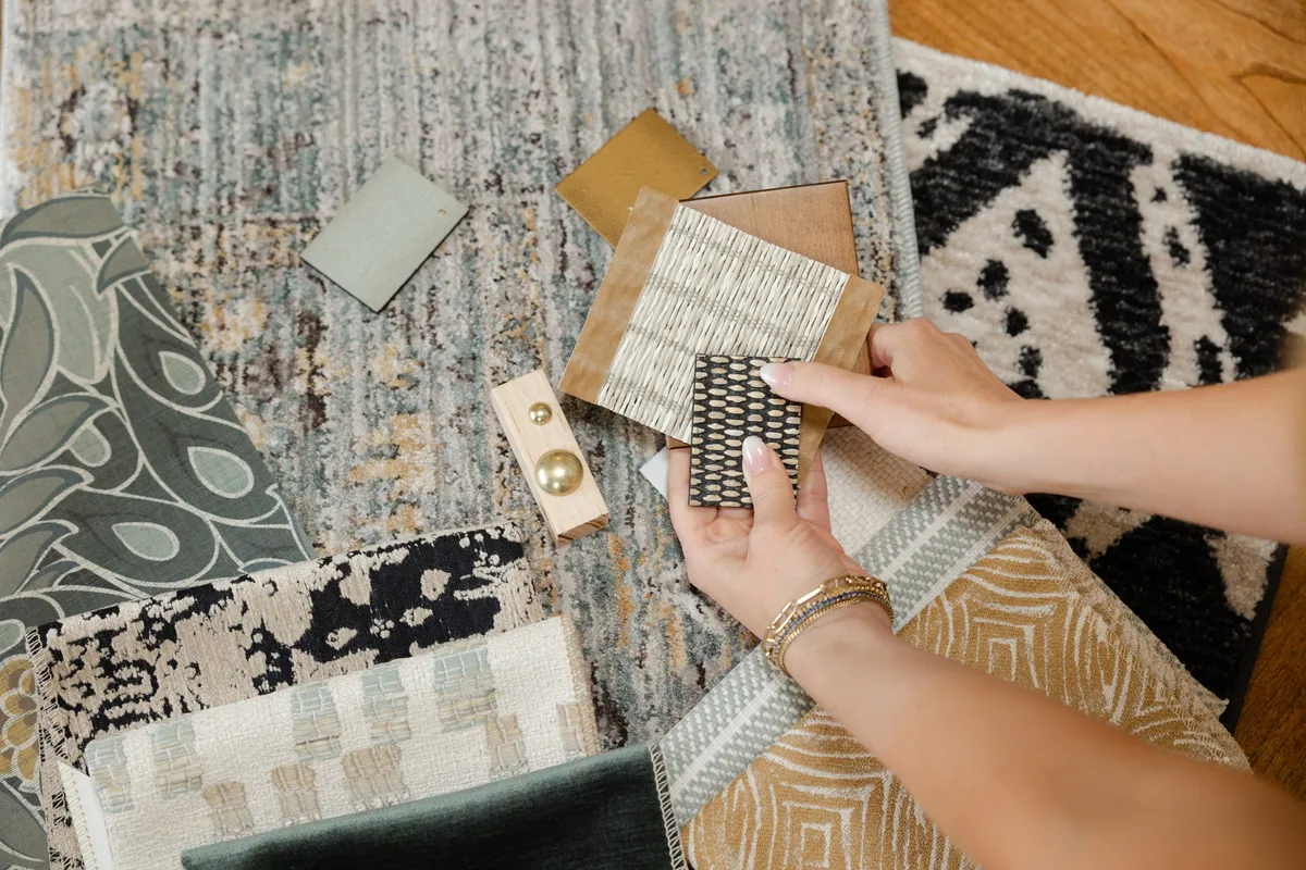 Hands comparing woven fabric and wood samples above patterned rugs and brass hardware