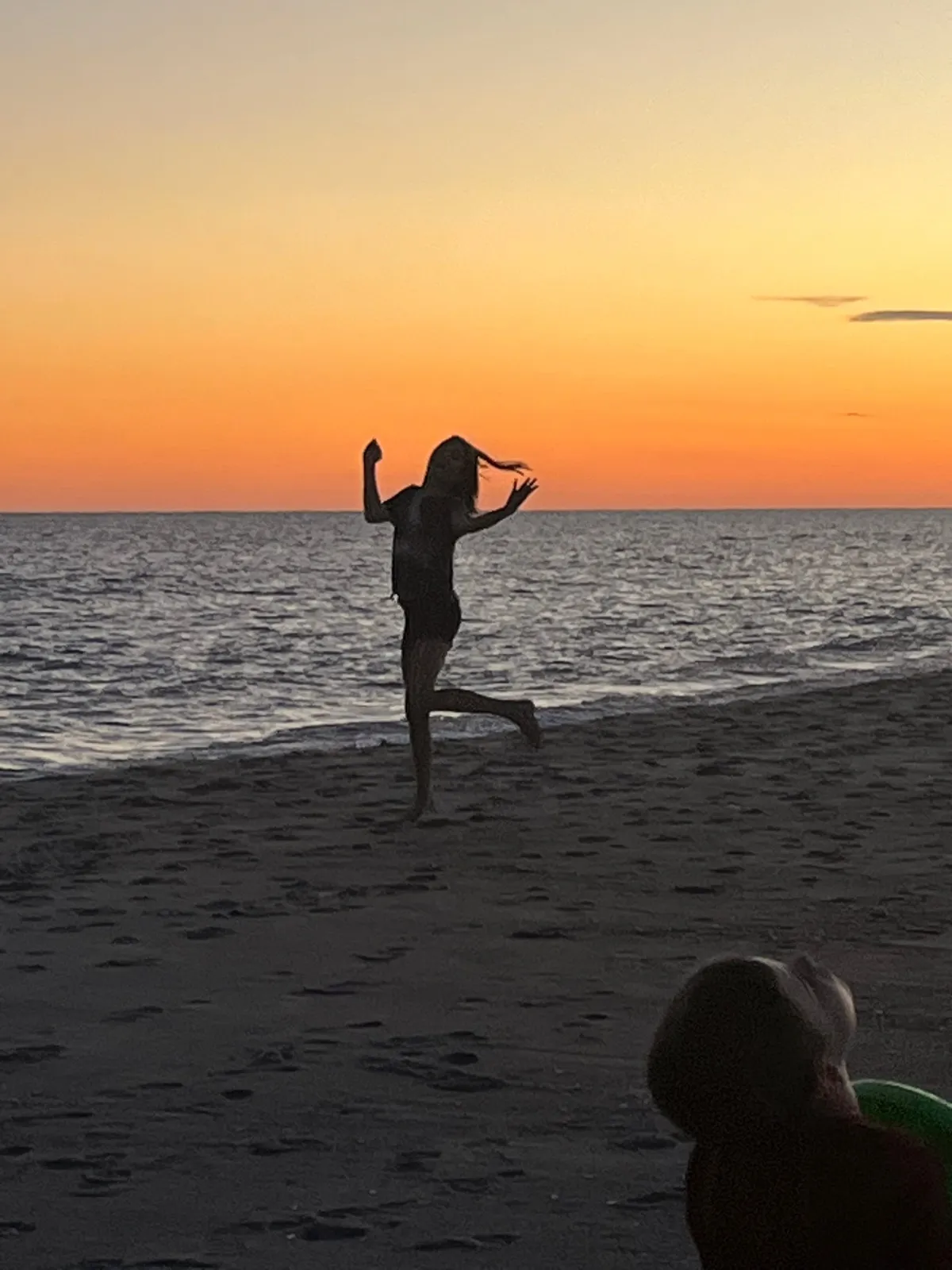 Silhouette of a person jumping on a sandy beach at sunset, with the orange sky and calm sea in the background.