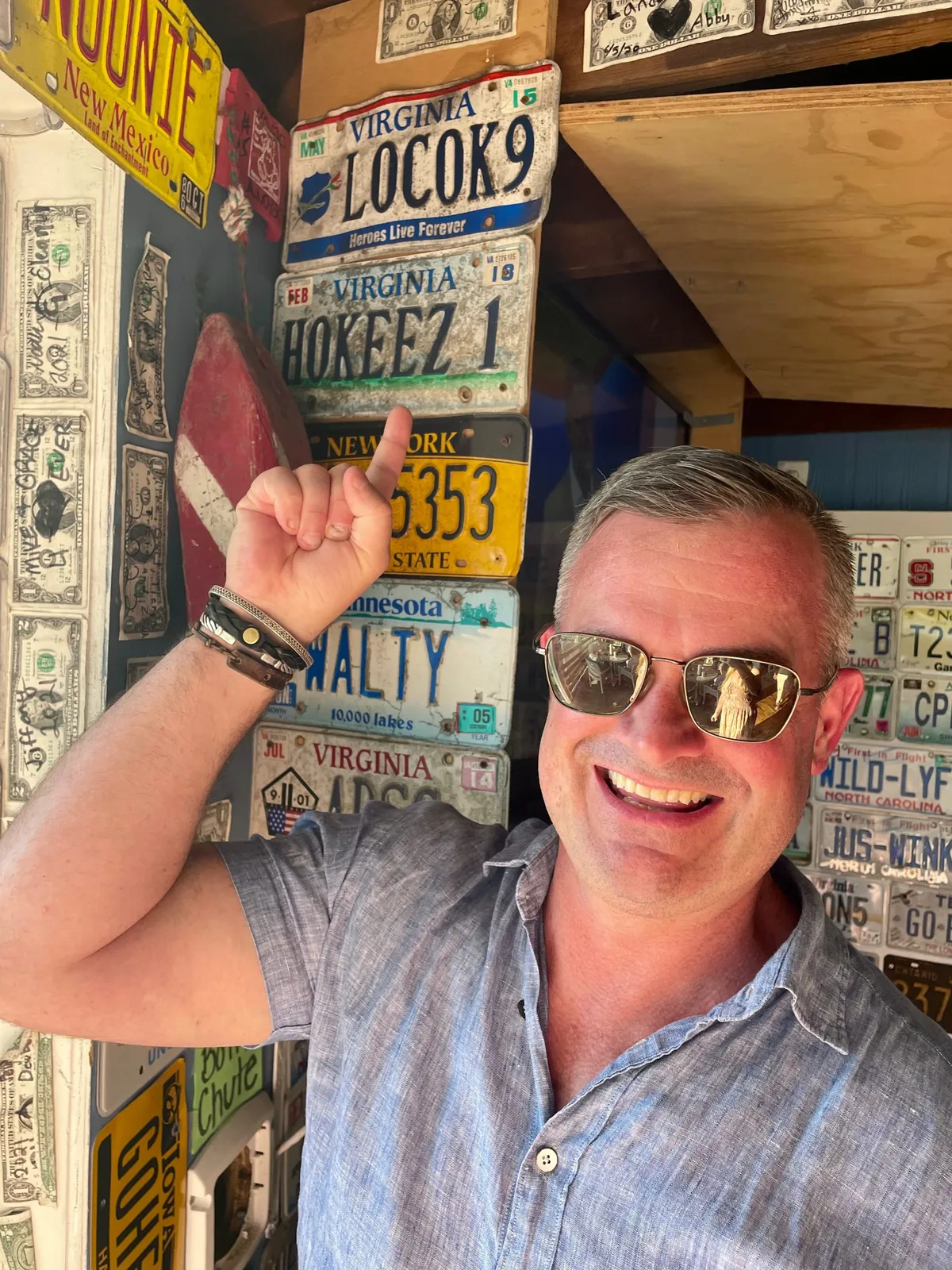Smiling man wearing sunglasses poses in front of a wall covered with Virginia license plates and vintage signs.