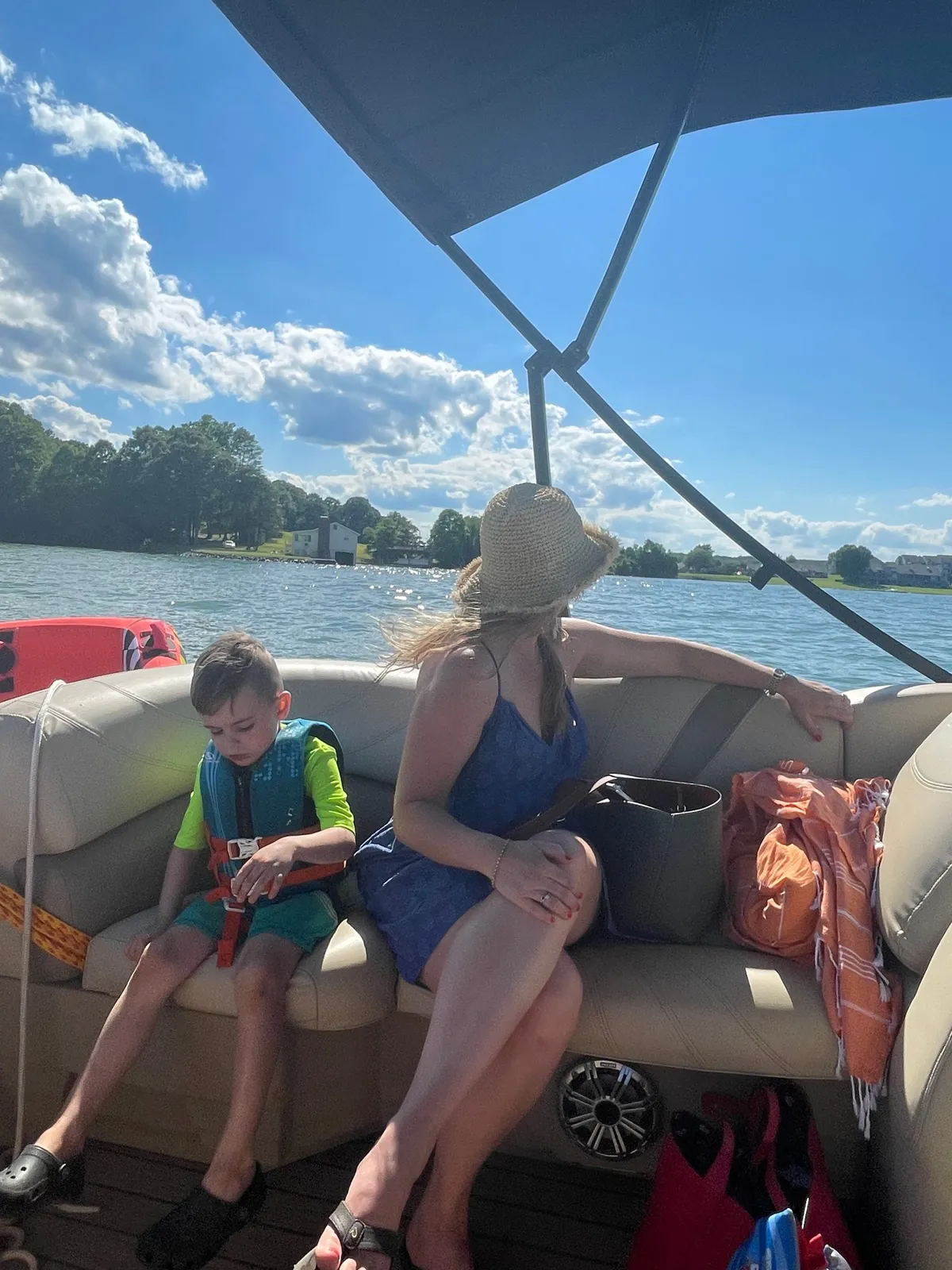 People riding in a boat on a lake: a woman in a straw hat and two children wearing life jackets sit on a cushioned seat with shoreline houses in the distance.