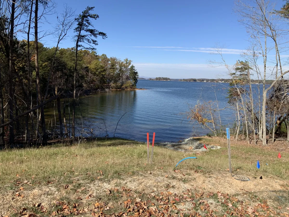 Lake view with calm water, forested shoreline, and autumn leaves on the grass; colorful stakes mark the foreground construction area.