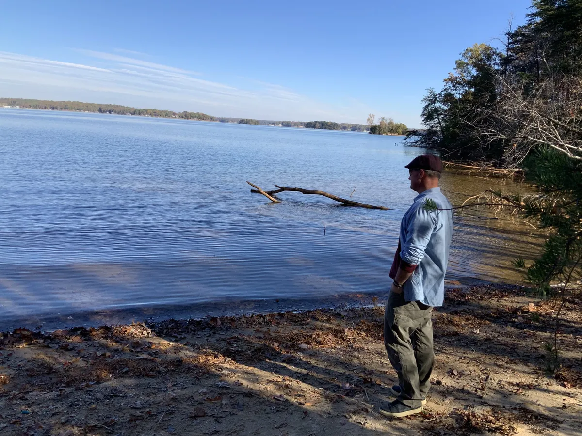 Man wearing a hat and light shirt stands at the water's edge on a sunny lakeshore with trees along the right bank.